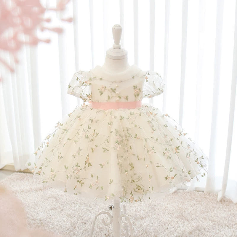 Un incantevole abito da principessa con un design etereo e raffinato. La gonna è realizzata con strati multipli di tessuto, creando un effetto voluminoso che dona leggerezza e movimento, come se fluttuasse nell’aria. L’ultimo strato, in tulle bianco trasparente, è arricchito da delicati ricami floreali in una romantica sfumatura di rosa tenue e verde, aggiungendo un tocco di poesia e raffinatezza.

🎀 Dettaglio del Cinturino:
Un elegante cintura rosa cipria,
