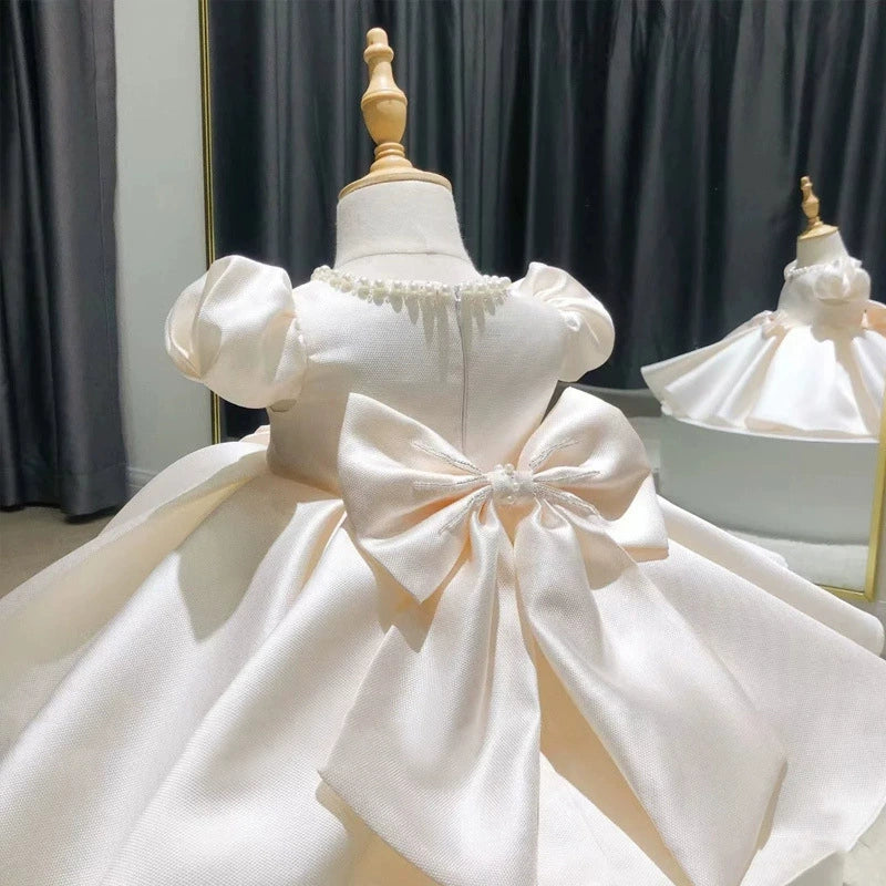 Un raffinato abito da cerimonia che incanta al primo sguardo! Realizzato in un tessuto satinato di alta qualità, questo vestito è perfetto per occasioni speciali come battesimi, prime comunioni e matrimoni. La gonna ampia e vaporosa è composta da più strati di tessuto, creando un effetto principesco e voluminoso che dona eleganza e movimento. Le maniche corte donano un tocco delicato e raffinato, mentre il cinturino in nastro di raso, finemente annodato sul davanti, aggiunge un dettaglio romantico e sofisti