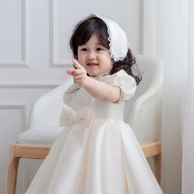 Nella seconda immagine, una dolcissima bambina di meno di due anni indossa questo incantevole abito, trasformandosi in una piccola principessa. Il tessuto satinato risplende alla luce, mettendo in evidenza la qualità del materiale e la cura nei dettagli sartoriali. La gonna, ampia e leggera, crea un effetto fluttuante che regala un’eleganza senza tempo. Il look è impreziosito da una graziosa collana di perle, che dona un tocco di classe, e dalla cintura in nastro, perfettamente abbinata al design dell’abito